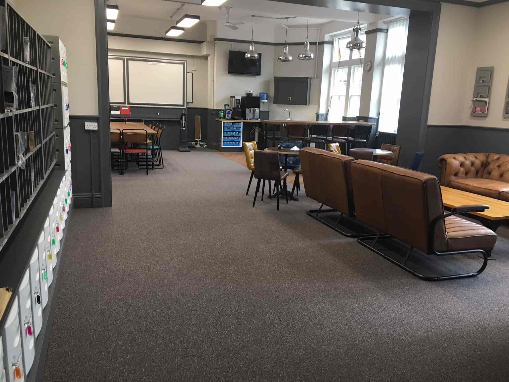 A spacious lounge area with brown leather sofas, a wooden coffee table, and high tables with stools. There are shelves with labeled files on the left and large windows letting in natural light.