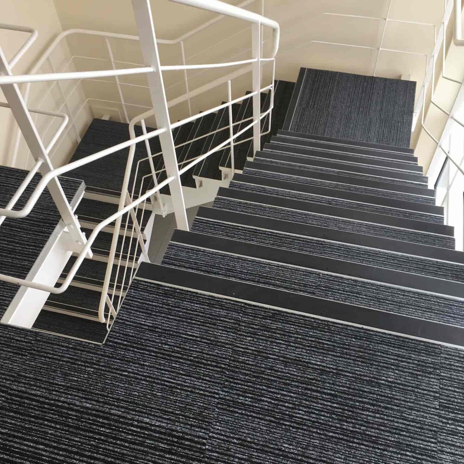A modern staircase with dark, striped carpeting and white metal railings, viewed from above. The stairs descend in two flights, turning at a landing. The walls and surroundings are light-colored.