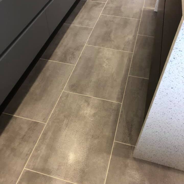 A close-up view of a kitchen floor with large, rectangular gray tiles and light-colored grout lines next to dark cabinets and a light, speckled countertop.