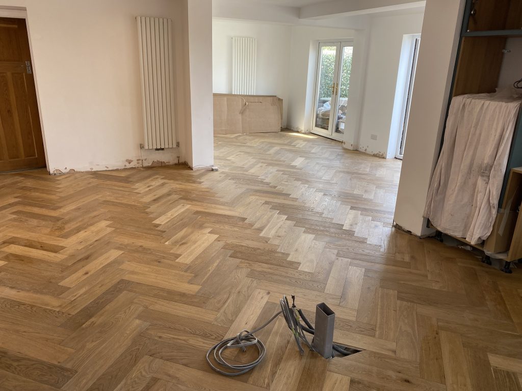 A room with newly installed herringbone wood flooring, unfinished baseboards, some exposed cables in the center, a covered cabinet, and double glass doors leading outside. Natural light enters through the doors and windows.