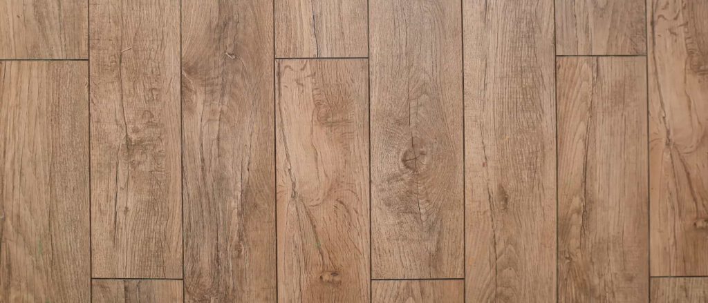 Wooden floor with long, light brown planks arranged in parallel rows. The planks have visible wood grain patterns, knots, and subtle variations in color, giving a natural, rustic appearance.