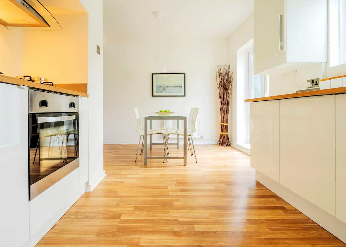 Bright, modern kitchen and dining area with wooden floors, white cabinets, a stainless steel oven, a small dining table with four chairs, a framed picture on the wall, and a tall vase with decorative branches.