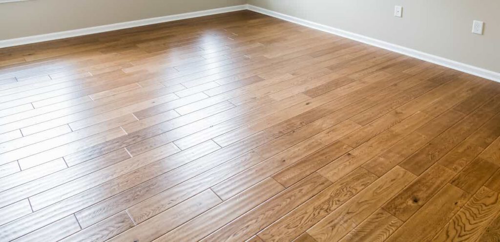 Empty room with light brown hardwood flooring, beige walls, white baseboards, and two electrical outlets visible on the right wall. Sunlight is reflecting off the floor near the windows.