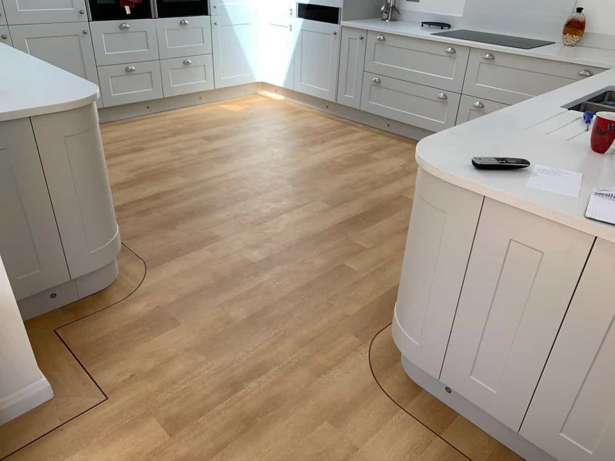 Modern kitchen with light wood flooring, white cabinets, and curved edges on the counters. On the right counter are a red mug, remote control, and notepad. Sunlight streams in from the back.