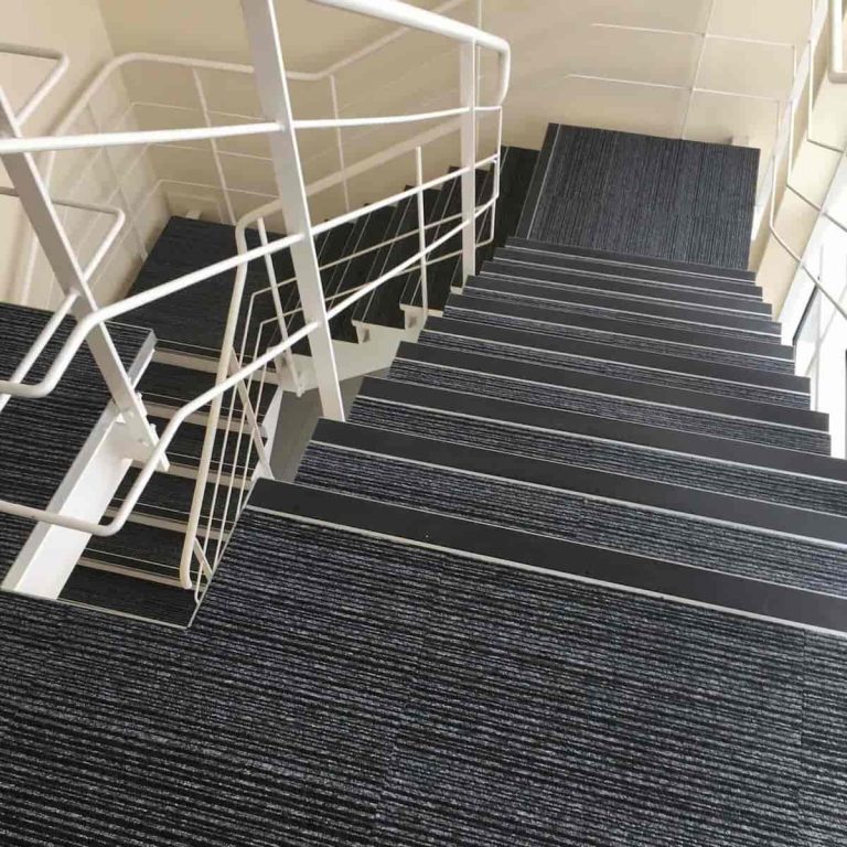 A modern indoor staircase with dark, striped carpeting and white metal railings, shown from the top looking down across several flights. The walls and railings are light-colored, creating a clean, minimalist look.