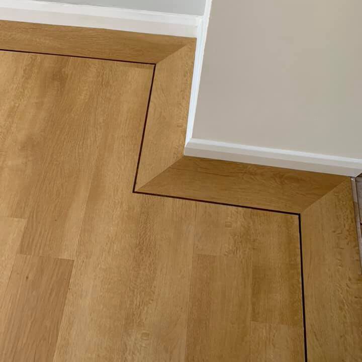 Wooden flooring with a darker border outline forming a geometric, zigzag pattern around the corner of a cream-colored wall and white skirting board. The detail highlights the precision of the flooring installation.